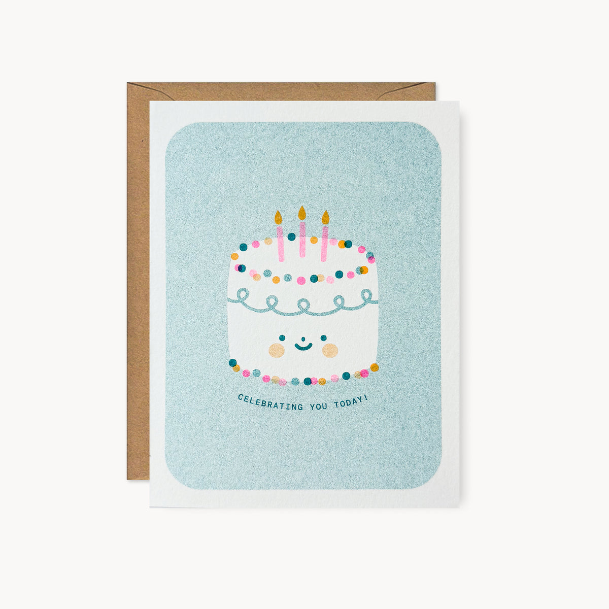 Whimsical risograph birthday card printed with teal, pink, and yellow vegetable-based inks, featuring a cute smiling frosted cake with rosy cheeks, three pink candles, and colorful dotted decorations on a teal background. The words "Celebrating You Today!" curve beneath it, and a kraft envelope is included. A delightful handmade card from Seattle, WA by Minna May.