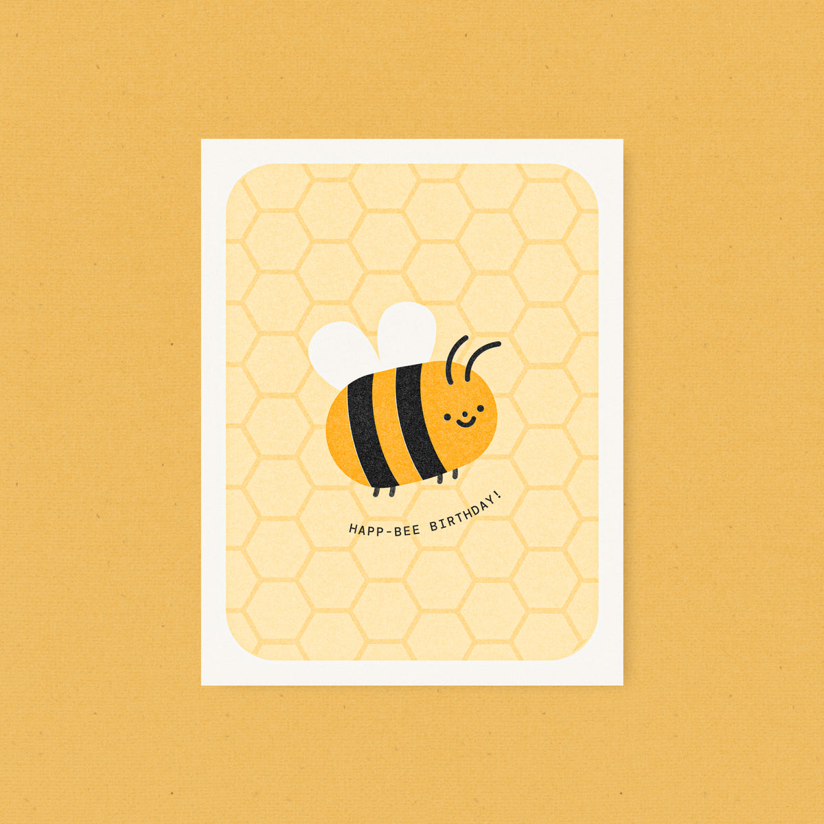 Whimsical risograph birthday card printed with yellow and black vegetable-based inks, featuring a cute smiling bumblebee with white wings on a honeycomb pattern. The words "Happ-Bee Birthday!" curve beneath it, and a kraft envelope is included. A delightful handmade card from Seattle, WA by Minna May.