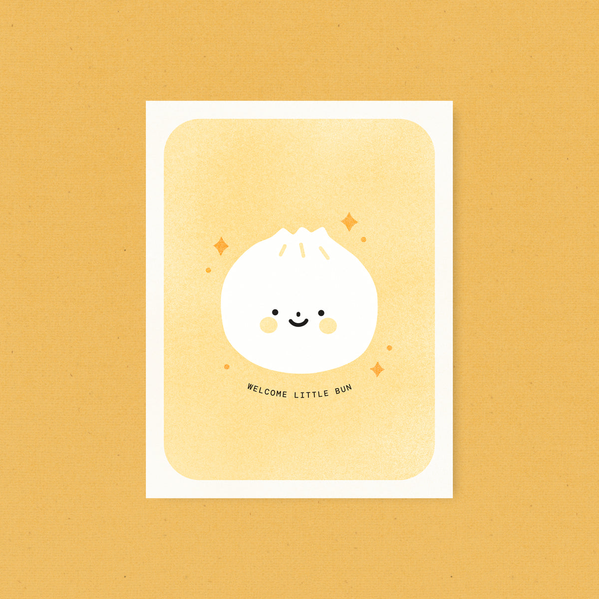 Whimsical Asian food-inspired risograph new baby card printed with yellow and black vegetable-based inks, featuring a cute smiling dumpling bun with rosy cheeks and little orange sparkles on a peach background. The words "Welcome Little Bun" curve beneath it, and a kraft envelope is included. A delightful handmade card from Seattle, WA by Minna May.