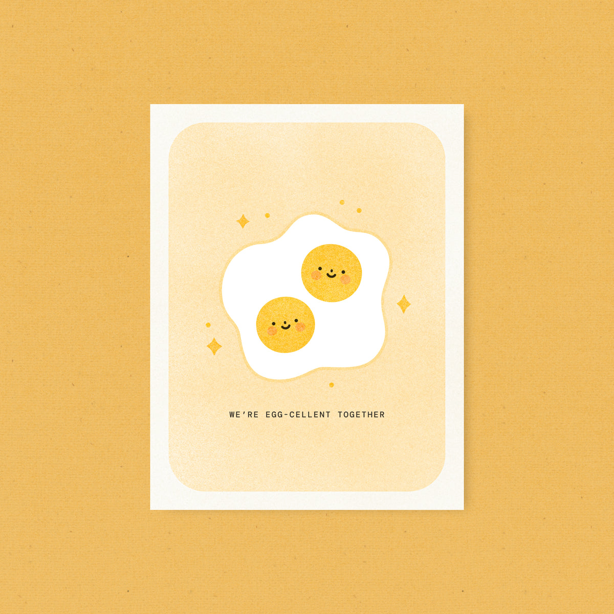 Whimsical risograph love card printed with yellow vegetable-based ink, featuring a cute fried sunny side up egg with two smiling yolks and rosy cheeks, surrounded by little sparkles. The words "We're Egg-cellent Together" appear below, and a kraft envelope is included. A delightful handmade card from Seattle, WA by Minna May.
