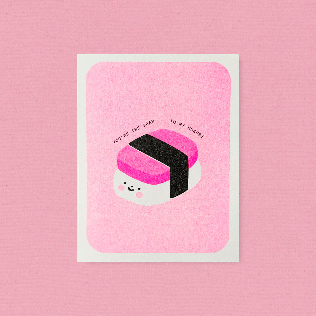 Whimsical Hawaii-inspired risograph greeting card printed with fluorescent pink vegetable-based ink, featuring a cute smiling Spam musubi with rosy cheeks and a black nori band. The words "You're the Spam to My Musubi" arch above it, and a kraft envelope is included. A delightful handmade card from Seattle, WA by Minna May Design.