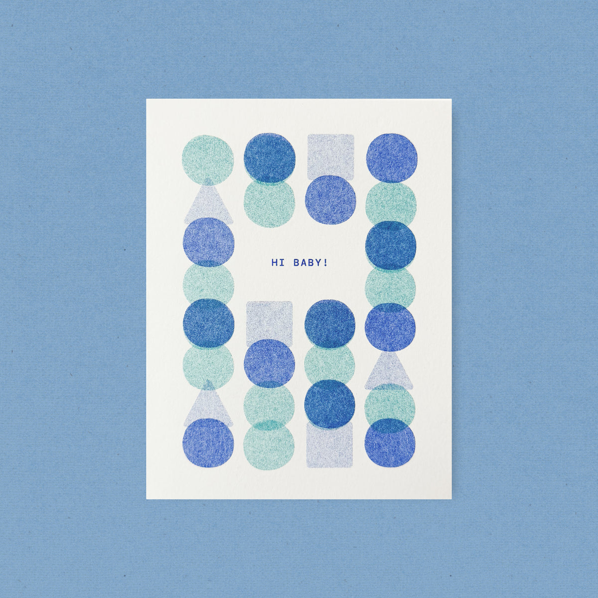 Whimsical risograph baby greeting card printed with sky blue and light teal vegetable-based inks, featuring a playful and modern pattern of overlapping blue, teal, and periwinkle circles, squares, and triangles. The words "Hi Baby!" sit in the center, and a kraft envelope is included. A delightful handmade new baby card from Seattle, WA by Minna May.
