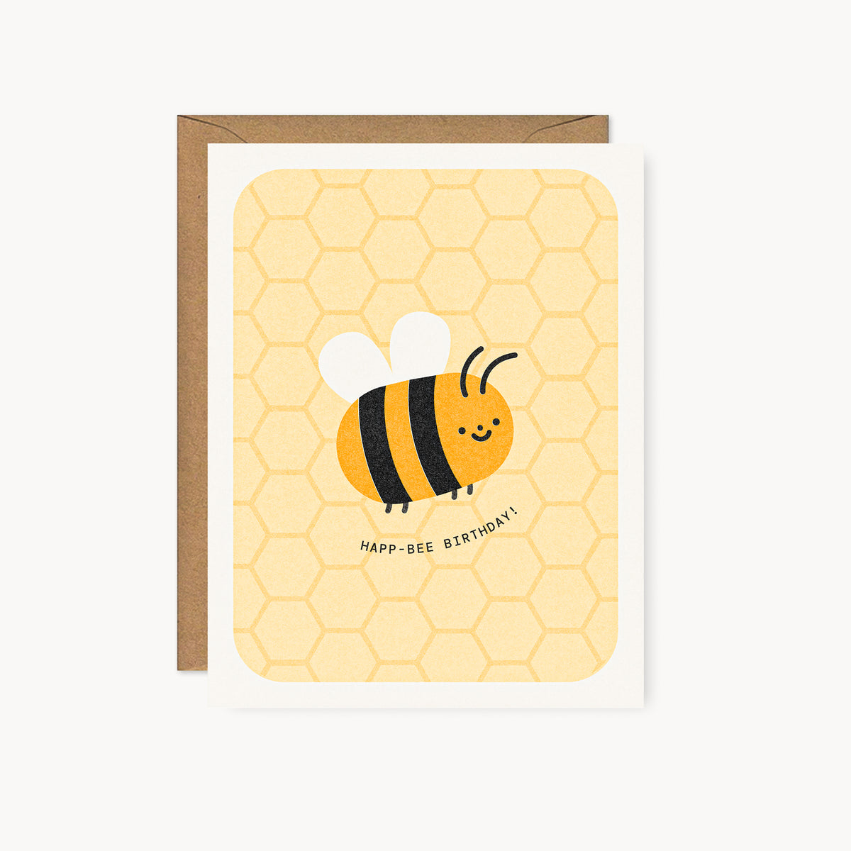 Whimsical risograph birthday card printed with yellow and black vegetable-based inks, featuring a cute smiling bumblebee with white wings on a honeycomb pattern. The words "Happ-Bee Birthday!" curve beneath it, and a kraft envelope is included. A delightful handmade card from Seattle, WA by Minna May.