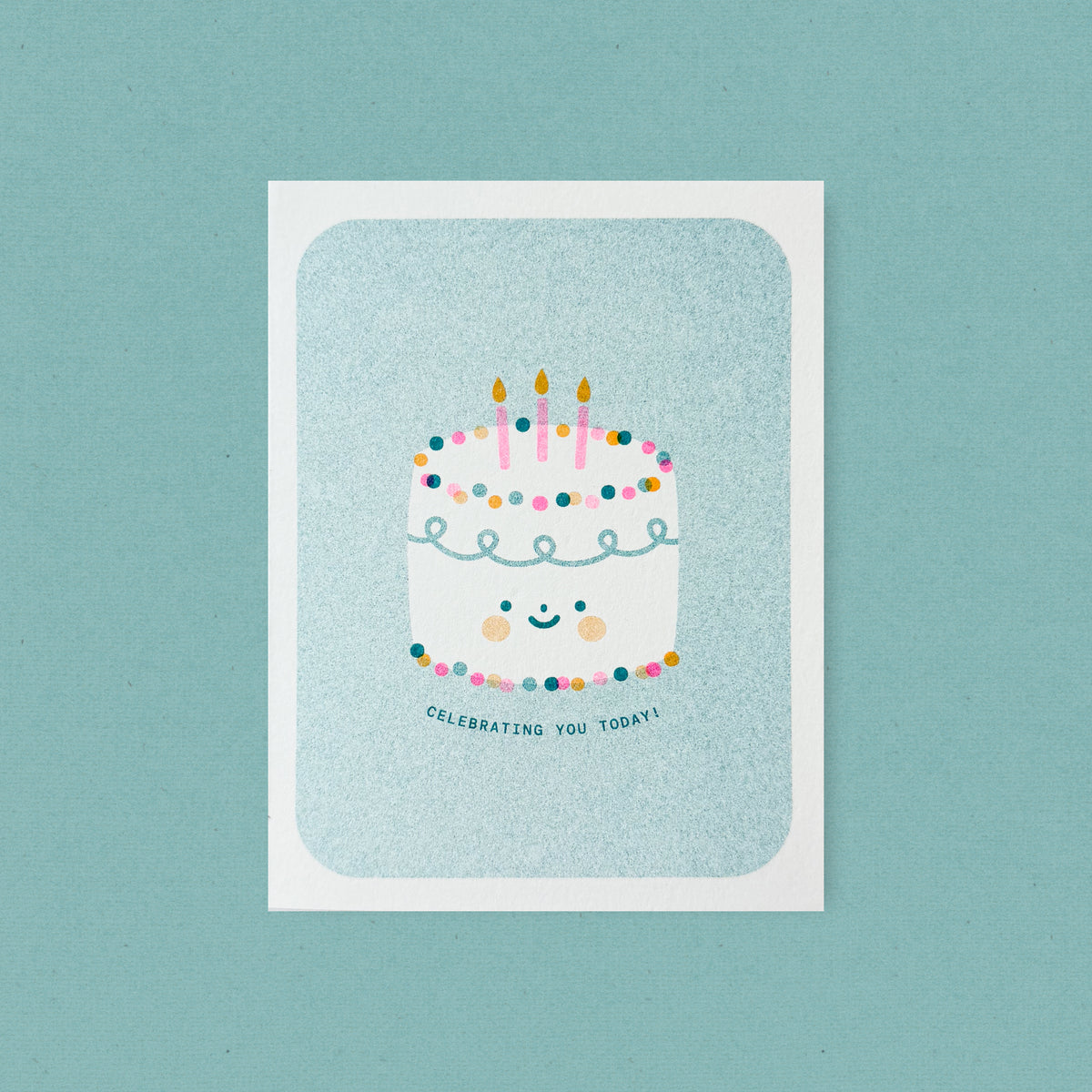 Whimsical risograph birthday card printed with teal, pink, and yellow vegetable-based inks, featuring a cute smiling frosted cake with rosy cheeks, three pink candles, and colorful dotted decorations on a teal background. The words "Celebrating You Today!" curve beneath it, and a kraft envelope is included. A delightful handmade card from Seattle, WA by Minna May.