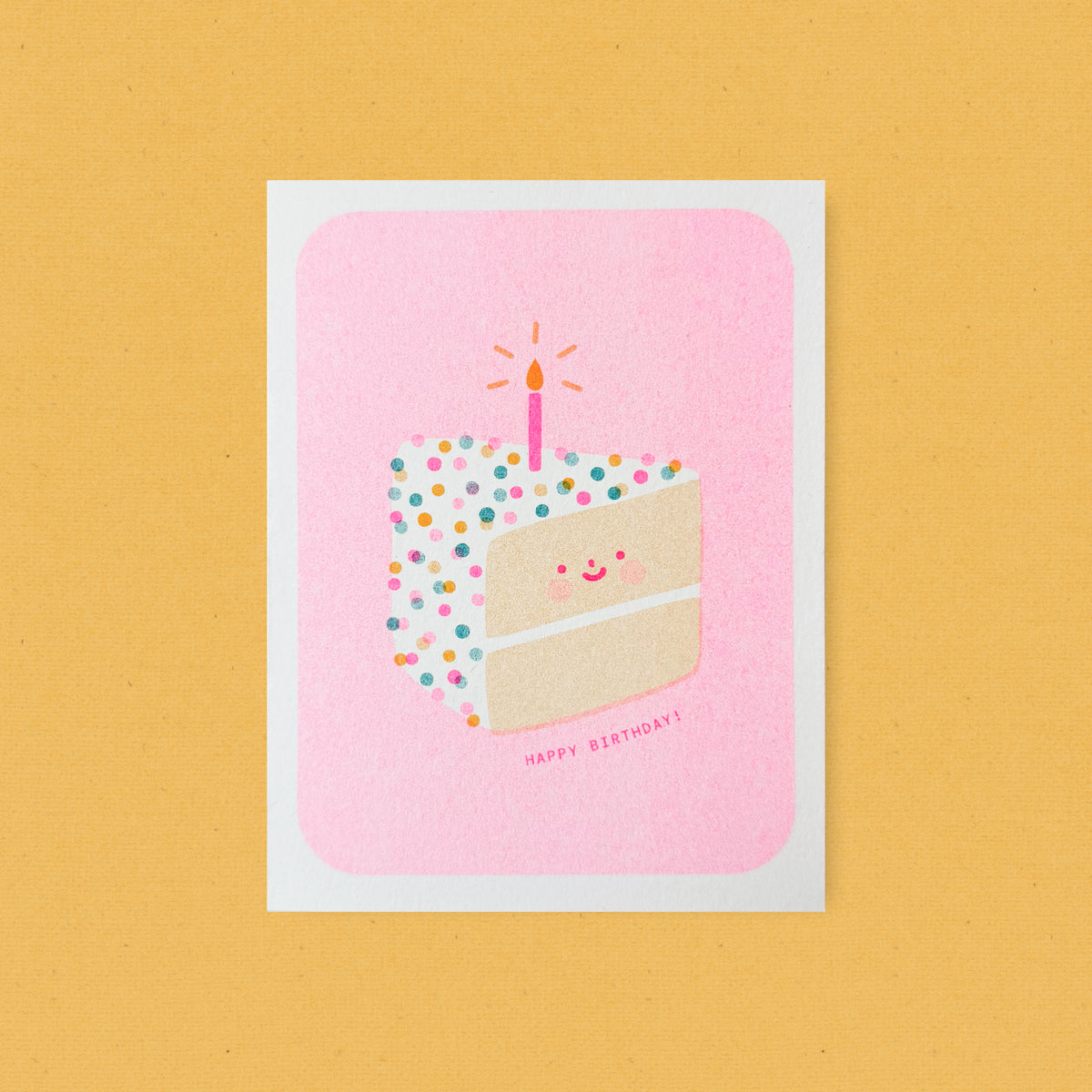 Whimsical risograph birthday card printed with fluorescent pink, yellow, and teal vegetable-based inks, featuring a cute smiling slice of confetti cake with rosy cheeks and a single glowing candle on a pink background. The words "Happy Birthday!" appear beneath it, and a kraft envelope is included. A delightful handmade card from Seattle, WA by Minna May.
