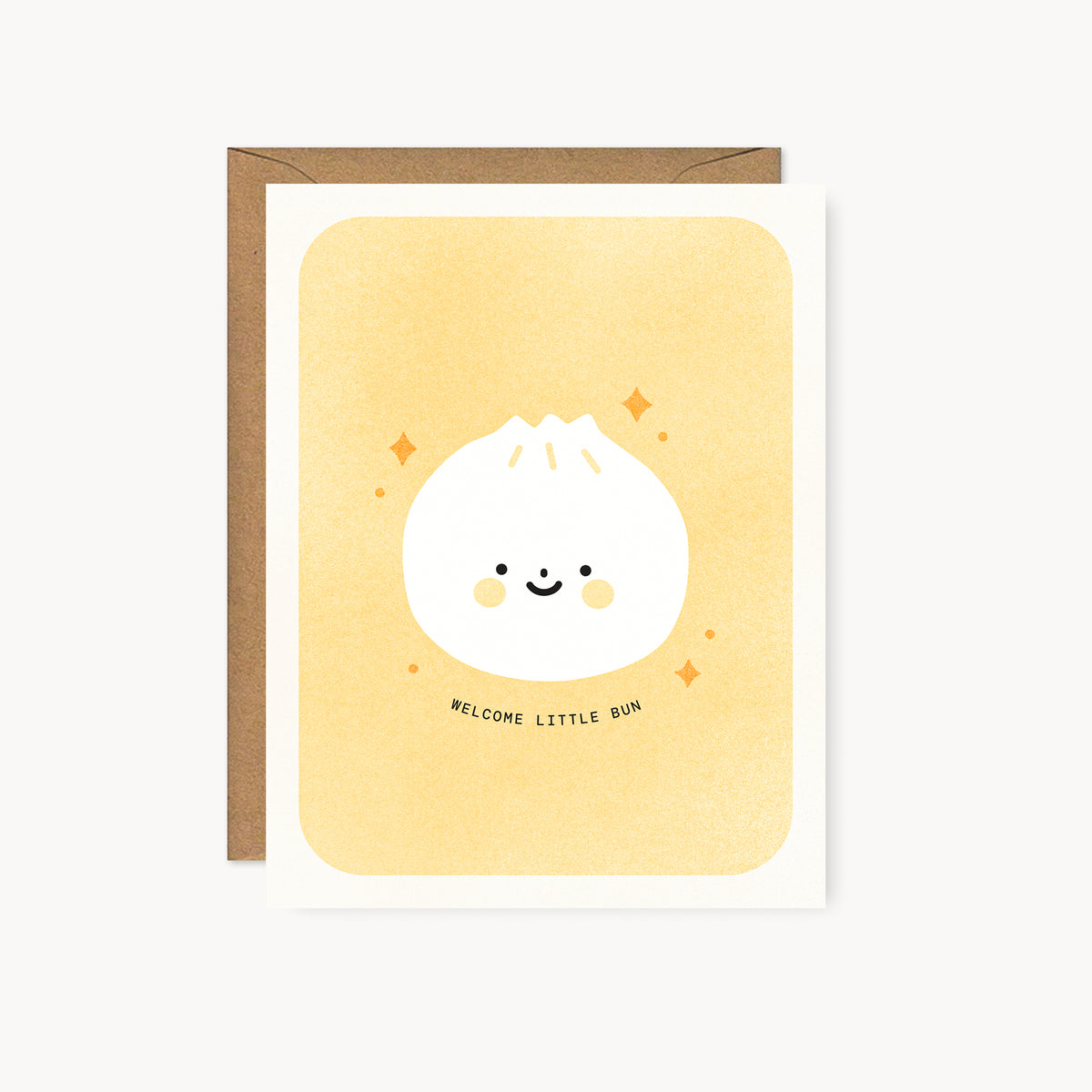 Whimsical Asian food-inspired risograph new baby card printed with yellow and black vegetable-based inks, featuring a cute smiling dumpling bun with rosy cheeks and little orange sparkles on a peach background. The words "Welcome Little Bun" curve beneath it, and a kraft envelope is included. A delightful handmade card from Seattle, WA by Minna May.